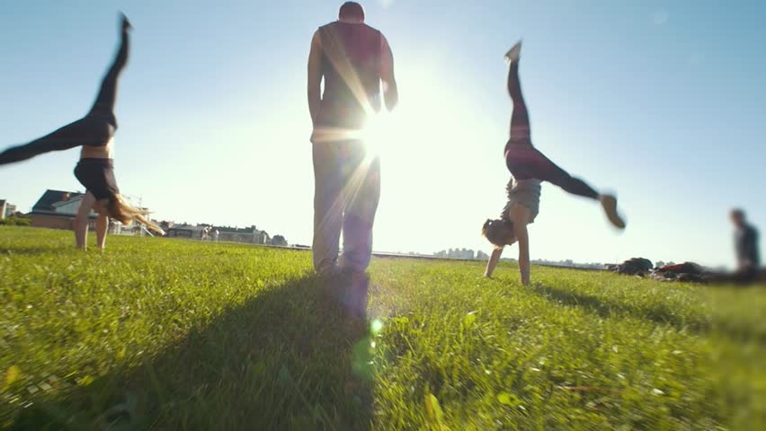 Young women performing acrobatic wheel on the grass, silhouette of young man standing in center of them | Shutterstock HD Video #1012547075