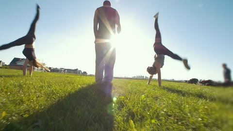 Young women performing acrobatic wheel on the grass, silhouette of young man standing in center of them