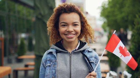 Slowmotion portrait of smiling African American girl traveller holding Canadian flag and looking at camera outdoors. Happy tourist and visiting foreign coutries concept.