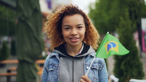 Slowmotion portrait of cheerful African American girl looking at camera and holding Brazilian flag standing in nice park in modern city. Tourism and people concept.