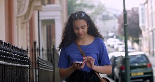 Portrait of Pretty Smiling Mixed Race Teen girl using her Smart Phone with Sunglasses, standing in front of London Street Housing. Curly Afro hair Beautiful Student Tourist exploring the British city.