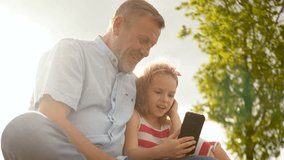 Grandfather and granddaughter look into the smartphone. Curly girl and mature man play games in mobile phone