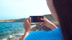 Rear view of brunette woman taking pictures of beautiful sea with smartphone. Girl making photo of rocky sea shore on sunny day on the beach. Hands holding smartphone. Summer vacation