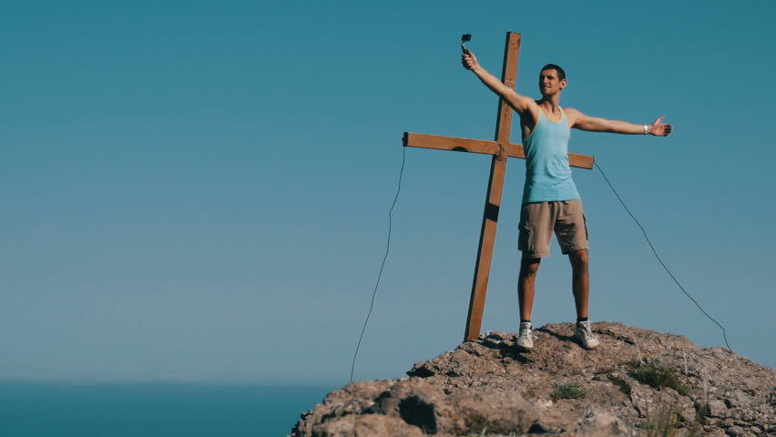 The young man joyfully takes himself off on the action camera, standing on top of the mountain near the cross. Achieving the goal, motivation