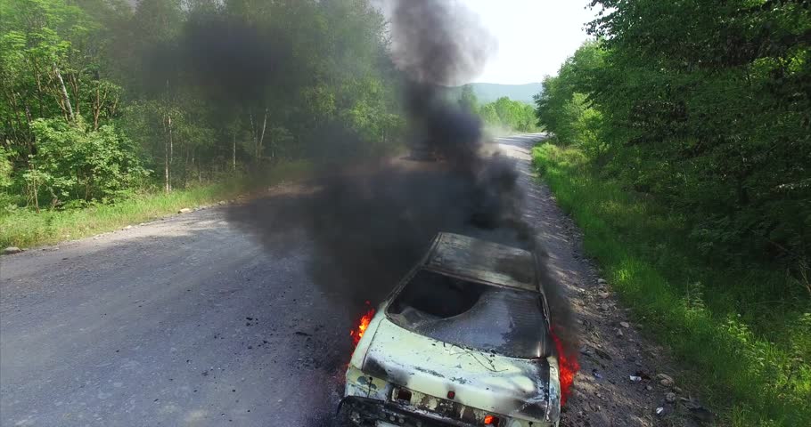 Aerial zooming out view of stolen car burning brightly with black smoke near road