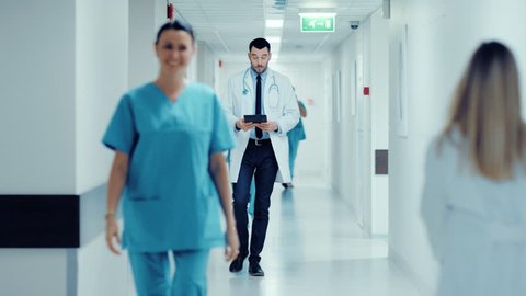 Determined Handsome Doctor Uses Digital Tablet Computer while Walking Through Hospital Hallway in Slow motion. Modern Bright Clinic with Professional Staff. Shot on RED EPIC-W 8K Helium Cinema Camera. Stockvideó