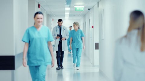 Surgeon and Female Doctor Walk Through Hospital Hallway, They Consult Digital Tablet Computer while Talking about Patient's Health. Modern Bright Hospital with Professional Staff. Shot on RED EPIC-W 8