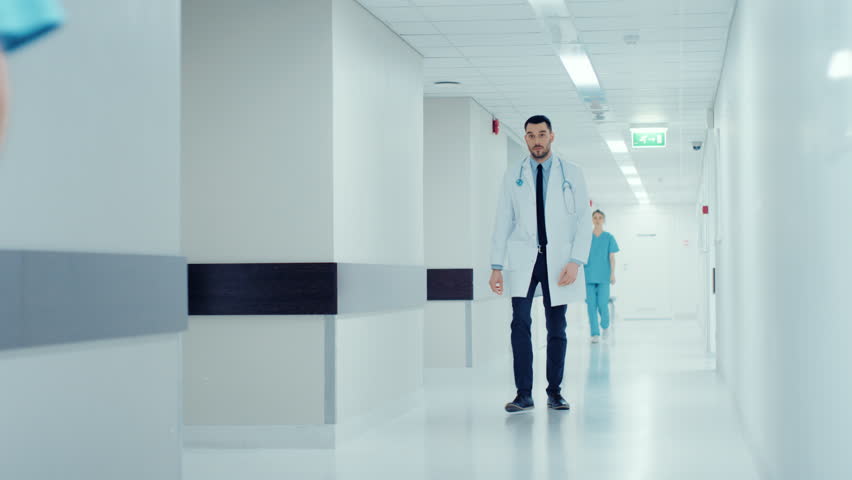 Determined Handsome Doctor Wearing White Coat with Stethoscope Walks Through Hospital Corridor in Slow motion. Modern Bright Clinic with Professional Staff. Shot on RED EPIC-W 8K Helium Cinema Camera.