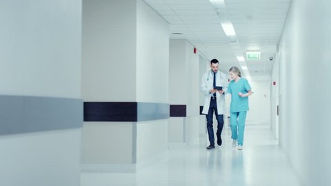 Surgeon and Female Doctor Walk Through Hospital Hallway, They Consult Digital Tablet Computer while Talking about Patient's Health. Modern Bright Hospital with Professional Staff.Shot on RED EPIC-W 8K