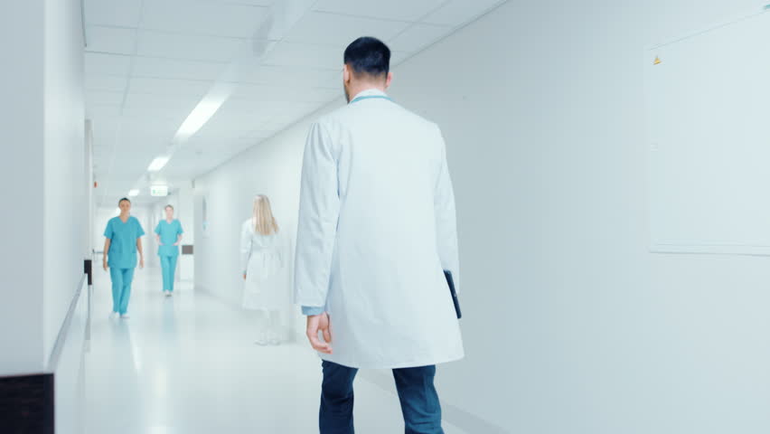 Following Back View Shot of a Doctor in a Hurry Walking Through Hospital Hallway. Greeting Nurses and His Colleagues. Bright Modern New Clinic. Shot on RED EPIC-W 8K Helium Cinema Camera. Royalty-Free Stock Footage #1012593926
