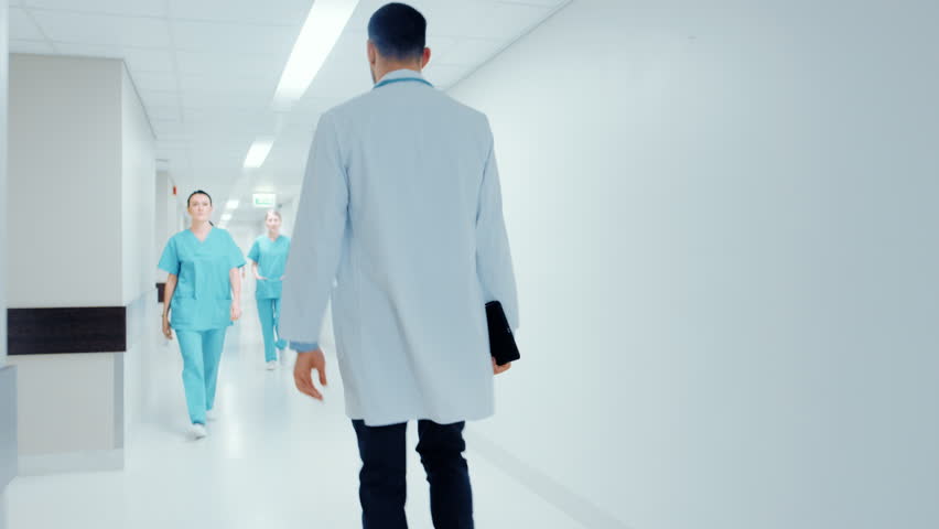 Following Back View Shot of a Doctor in a Hurry Walking Through Hospital Hallway. Greeting Nurses and His Colleagues. Bright Modern New Clinic. Shot on RED EPIC-W 8K Helium Cinema Camera.