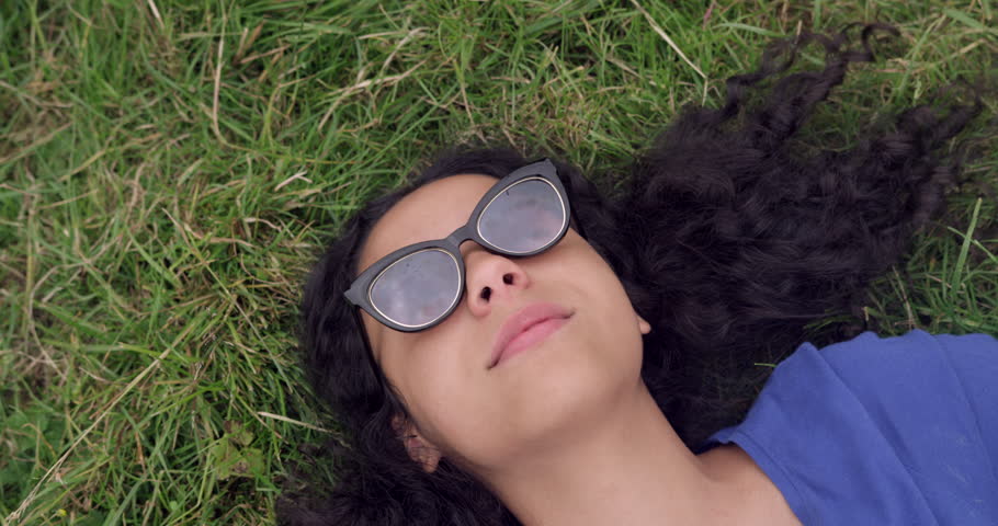 Portrait of Happy Beautiful Mixed Race Teen Girl Laying on green grass field smiling with sunglasses on. Curly afro haired student woman having fun in summer on gap year. 