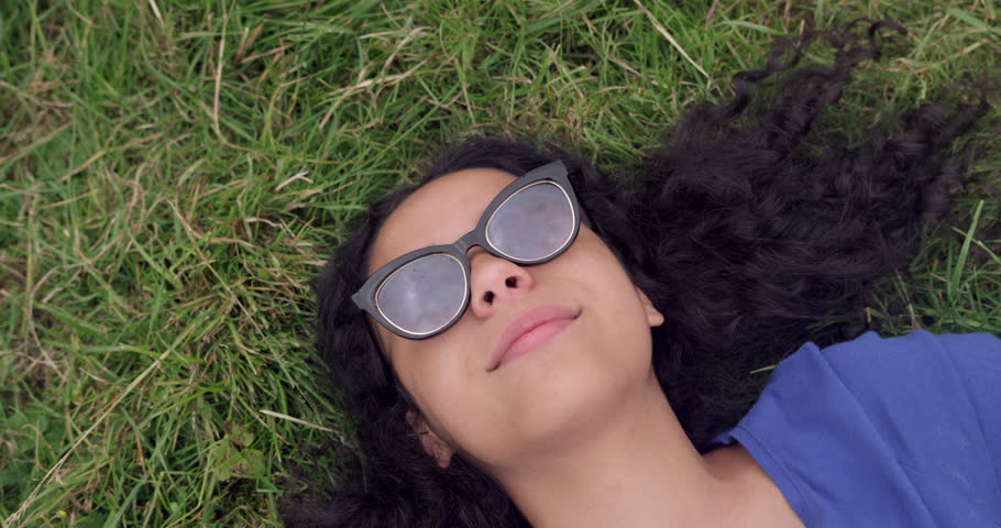 Pretty mixed race woman laying on grass with sunglasses on. Cute girl with big afro hair smiling shot from above. Summer time on field playing in the park. Student Taking glasses off to say hello. 