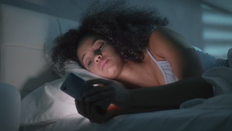 Young latino woman in bed messaging with mobile telephone at home. Tired african american girl in bedroom with cell phone. People using smartphone for texting message before sleeping