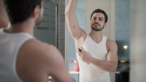 Young hispanic people and male beauty. Confident metrosexual man using spray deodorant on underarm skin, smiling and looking at mirror. Slow motion