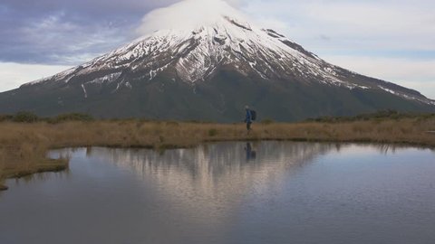 Slow motion - traveler wlking cross view to the taranaki volcano in the north island of new zealand and with the reflection of tho mountain and the climber in a lake. New Zeland