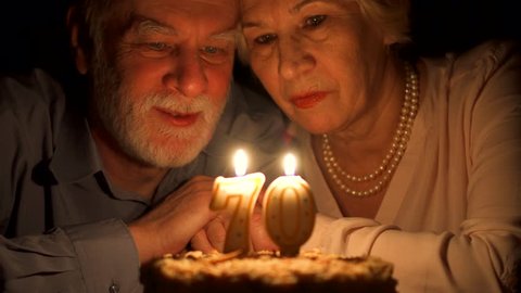 Loving senior couple celebrating anniversary with cake at home in evening. Happy elderly family hugging, cuddling together, make wishes and blowing out candles in form of number 70. Focus on seniors