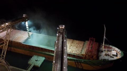 Night panorama of loading grain crops on bulk freighter ship via trunk to open cargo holds at silo terminal in seaport from above. Cereals bulk transshipment. Transportation agricultural products.
