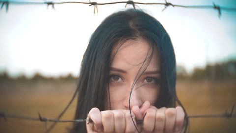 girl brunette refugee behind barbed lifestyle wire camp slow motion video. the concept of freedom is upset Woman hands and barbed wire. girl Refugee, prison, refugees captivity concept