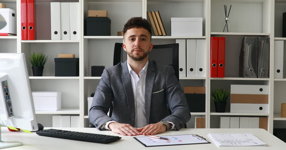 businessman in gray jacket sitting at table in white office and looking at camera