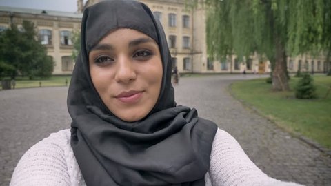 Young happy muslim girl in hijab is making video, saying hey, waving hand in blue weather, communication concept, religious concept, building on background