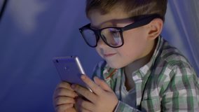 internet child in eyeglasses watches video into mobile phone in the wigwam close-up