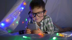 little smart boy with glasses watching video on mobile phone hiding in tent with garland at home close-up