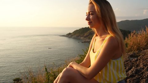 Slow motion medical student female relaxing after morning jogging, fair-haired lady sitting on rock enjoying beautiful view. Concept of tourism, summer clothes and travelling. Young smiling woman