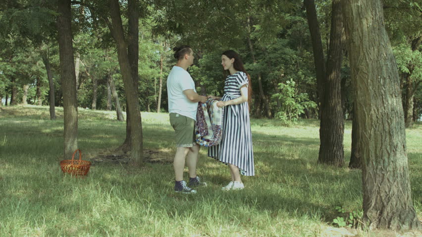Positive attractive pregnant couple in love putting picnic blanket on green grass in public park. Joyful young family spending leisure in nature and picnicking.