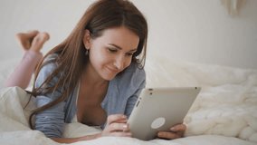 Beautiful woman with brown long hair lying on the bed with white sheets and communicating in social networks on her brand-new tablet. Indoors. Portrait.
