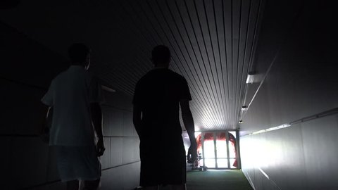 Two football players are walking along a dark tunnel to the football field. View from the back