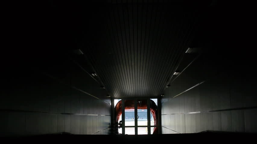 Two football players are walking along a dark tunnel to the football field. View from the back