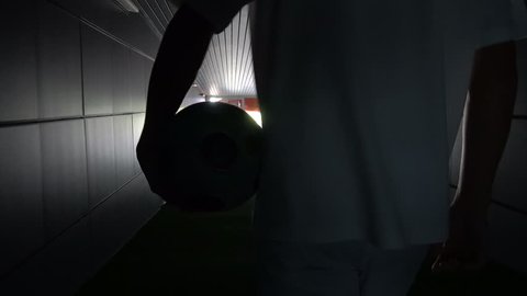 Two football players are walking along a dark tunnel to the football field. View from the back
