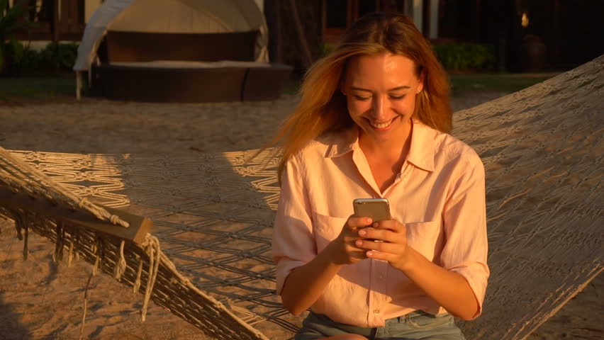 Slow motion HR manager lady holding smartphone sitting on hammock during vacation, girl smiling watching videos about dog stayed at home. Concept of innovative technologies, travelling and gadgets Royalty-Free Stock Footage #1012715243