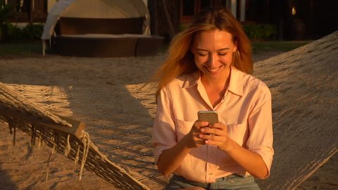 Slow motion HR manager lady holding smartphone sitting on hammock during vacation, girl smiling watching videos about dog stayed at home. Concept of innovative technologies, travelling and gadgets