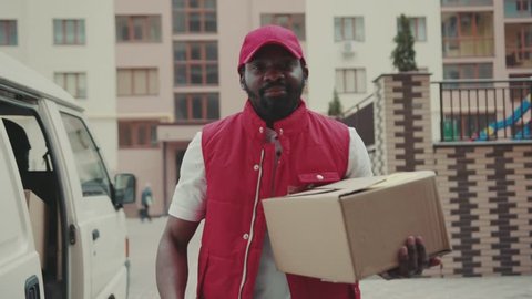 Portrait handsome courier young african american man with cap look at camera smile hold box delivery postal uniform job worker cardboard post distribution transport package logistic