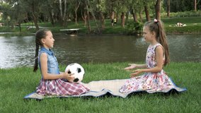 Two charming little girls sit at sunset in the Park on the lawn against the river and play with a soccer ball, throwing it to each other.