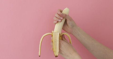 Female hands hold phallic fruit play and imitates jerk. Close up of peeled banana representing man penis while masturbating on pink background
