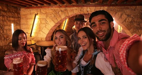 Young multi-ethnic friends in traditional German costumes celebrating Oktoberfest and taking selfies with beer mugs
