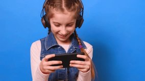 Little girl in headphones smiling and enthusiastically playing the video game on her smartphone on the blue background in studio. Young girl playing the video game and enjoys of victory.