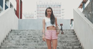 Young girl walks down stairs with skateboard. Sporty pretty woman with longboard winks to the camera. Back view. City style. 4K video shooting by handheld gimbal