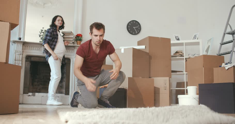 Future parents - cute man and a positive woman move in their new house and roll out the carpet in their new living room | Shutterstock HD Video #1012750247
