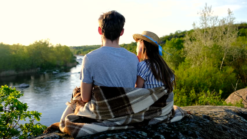 Loving couple wrapped in plaid blanket, sitting on mountain outdoors. Rear view. Man and woman travel together. Romantic traveler couple watching beautiful landscape, enjoying nature on sunset.