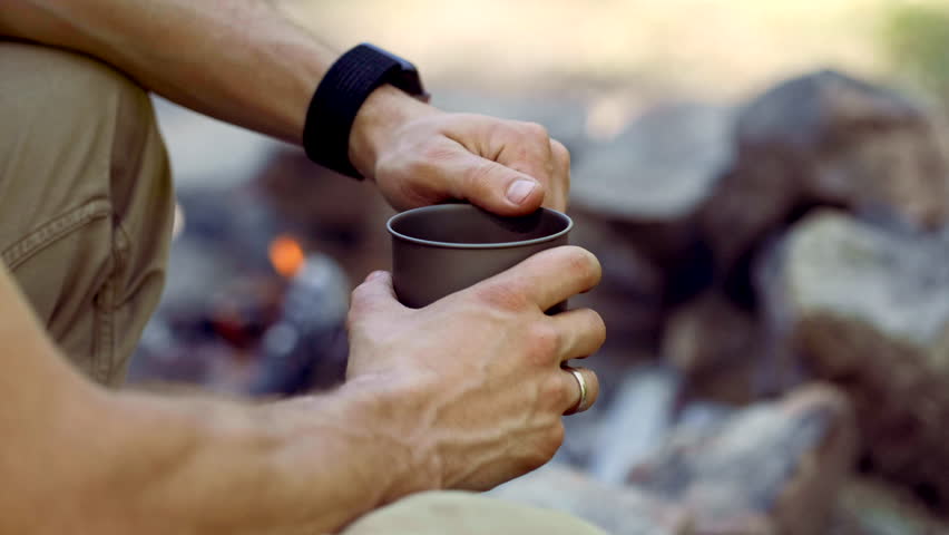 Man traveler hands holding cup of tea near the fire outdoors. Adventure, travel, tourism and camping concept. Hiker drinking tea from mug at camp. Coffee cooked over a campfire on the nature.