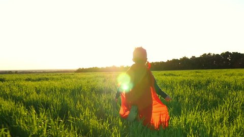 A happy child in a superhero costume in a red raincoat runs along a green lawn against the sunset, looking back.