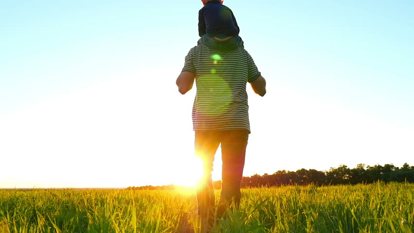 Happy young family walking on a green lawn, on a sunset background, in slow motion. Dad holds the child on his shoulders, Mom comes from behind