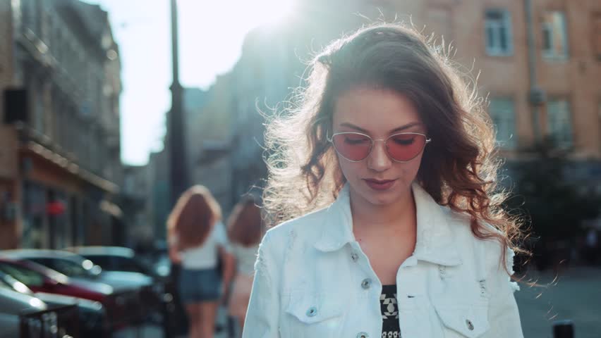 Smiling curly woman wearing trendy sunglasses walks down the central city street and uses her phone. Pretty summer woman in white jacket walks down the street looking at her mobile phone Royalty-Free Stock Footage #1012781630