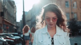 Smiling curly woman wearing trendy sunglasses walks down the central city street and uses her phone. Pretty summer woman in white jacket walks down the street looking at her mobile phone