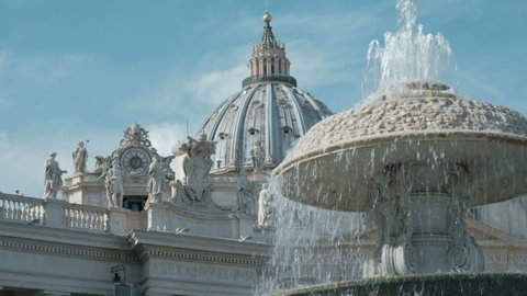 Low angle view camera tracking right past water fountain with view of the dome of St. Peter's Basilica in Vatican City