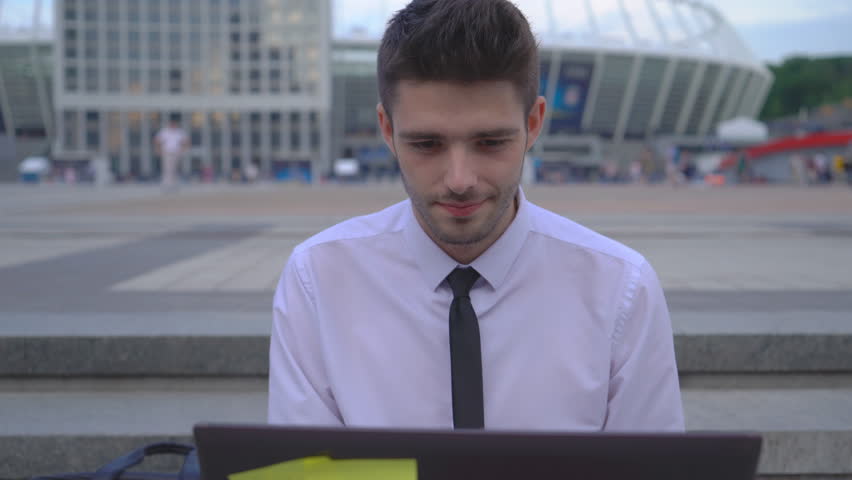 Young successful guy office worker working on the street on laptop. A man in a white shirt and tie. nice young man working on a project. | Shutterstock HD Video #1012794851